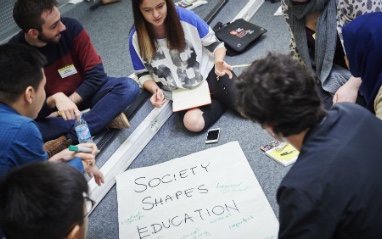 Fotografia di un gruppo di giovani seduti in cerchio per terra durante un laboratorio, attorno a un cartellone scritto a mano: «Society shapes education».