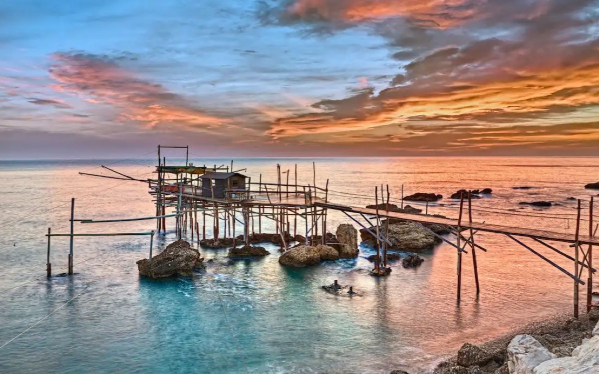 Fotografia di un trabocco sulla costa di San Vito Chietino al tramonto: la palafitta di legno con la casetta e le antenne per le reti, sopra un mare basso e trasparente.