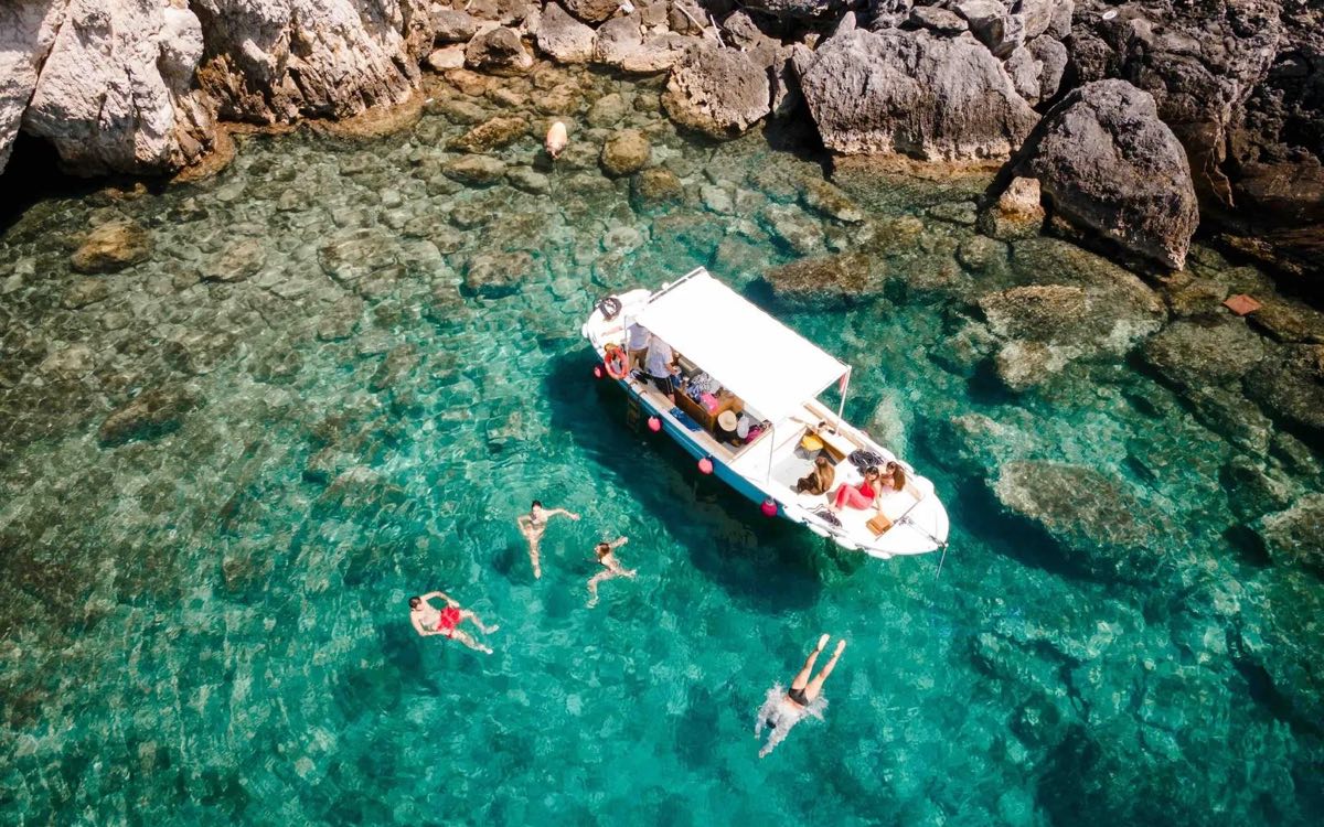 Fotografia dall'alto di una cala delle Isole Tremiti: una barca all'ancora nell'acqua turchese e trasparente, con alcune persone che nuotano intorno, sotto una costa di roccia bianca.